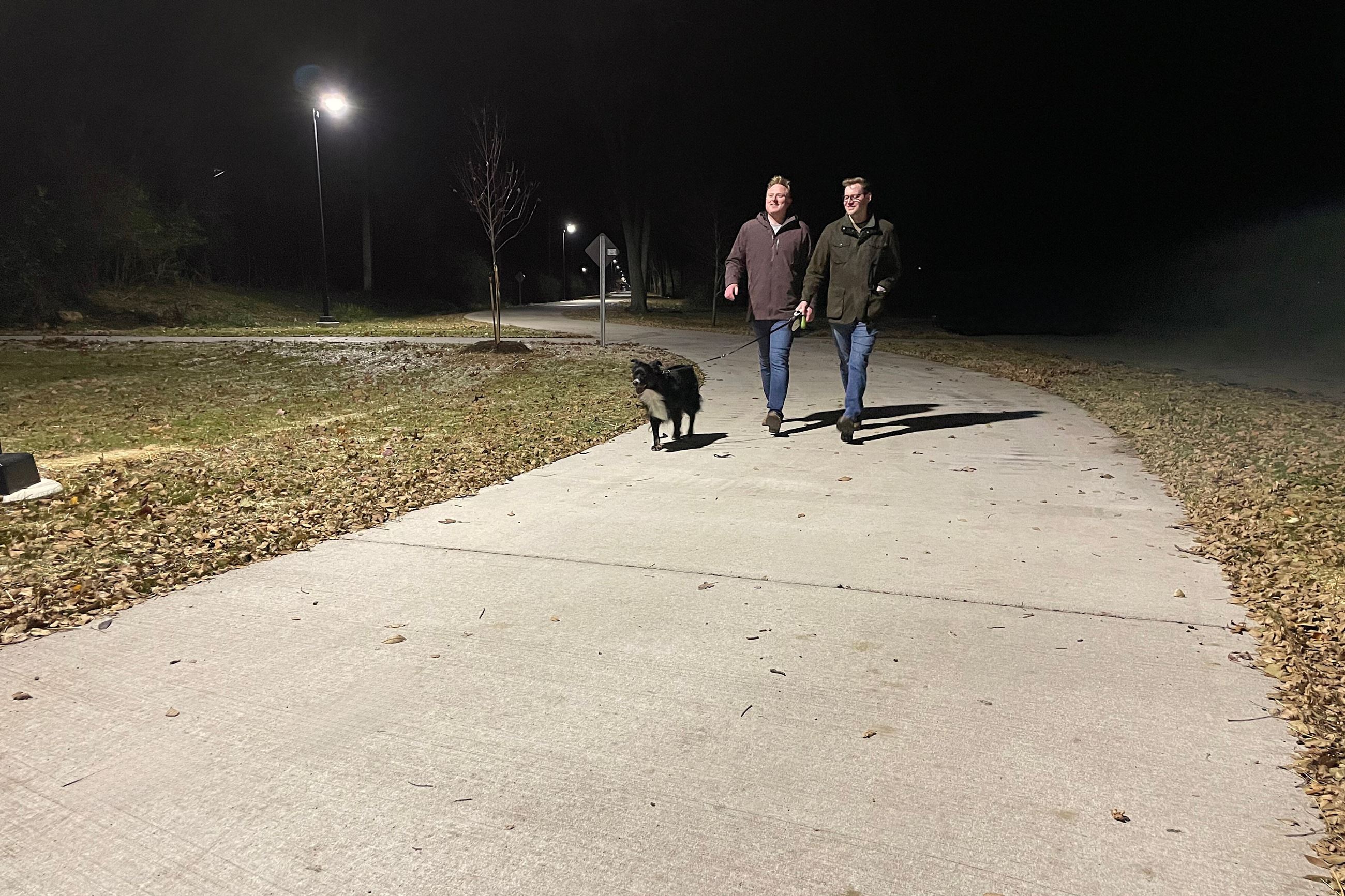 Two men walk dog on new concrete trail at night with bright lights overhead. 