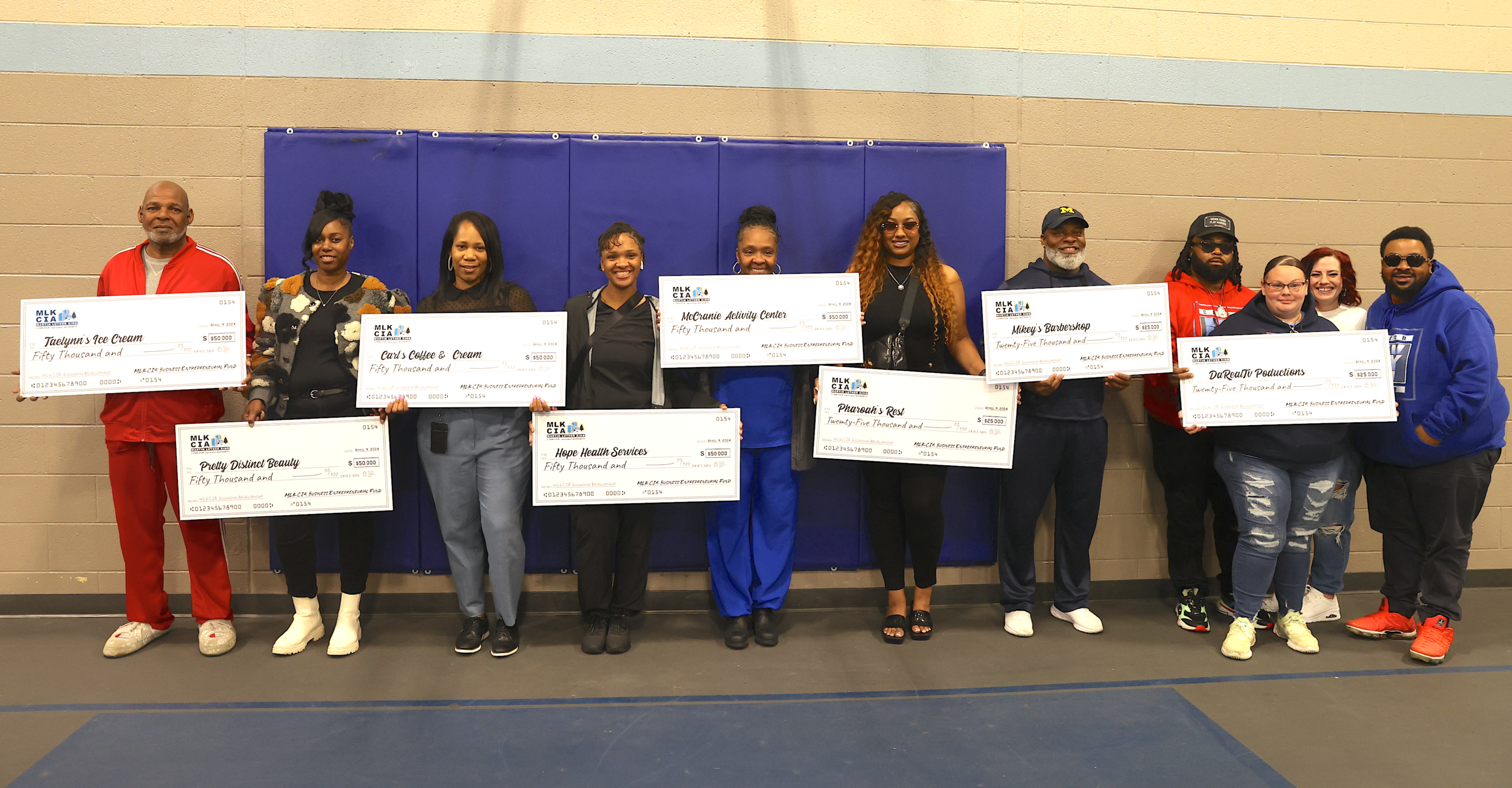 A line of people smiling in a gym holding oversized checks 